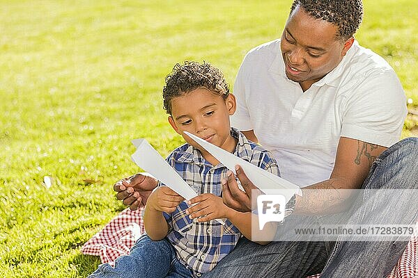 Glücklicher gemischtrassiger Vater und Sohn spielen mit Papierflugzeugen im Park