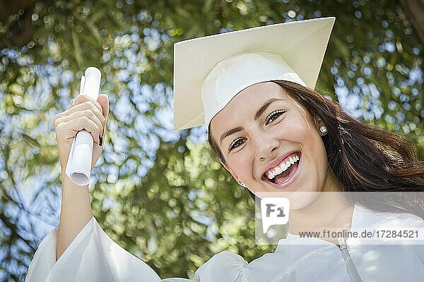 Attraktives gemischtrassiges Mädchen  das draußen mit Kappe und Talar mit Diplom in der Hand seinen Abschluß feiert
