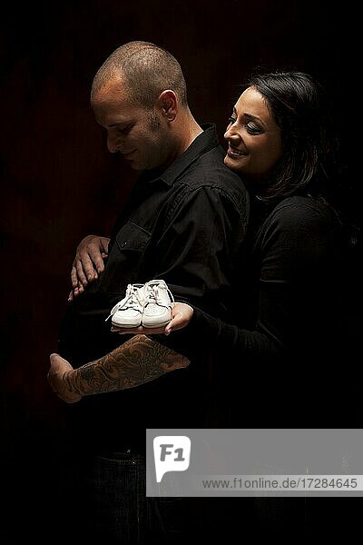 Fun gemischtrassiges Paar hält neue weiße Baby-Schuhe vor einem schwarzen Hintergrund unter dramatischen Beleuchtung