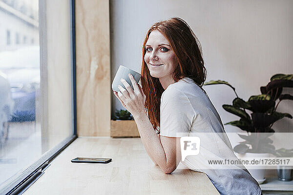 Beautiful woman having coffee leaning on window sill at home