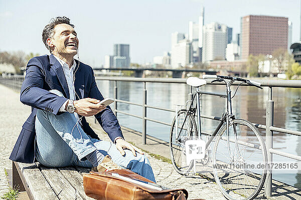 Happy mature businessman sitting by river during sunny