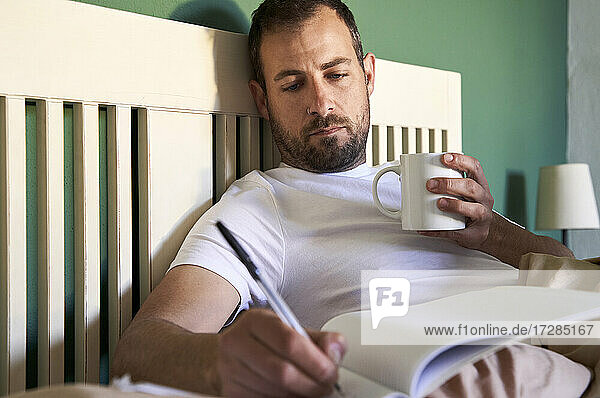 Mann mit Kaffeetasse schreibt in Buch bei der Arbeit zu Hause