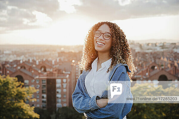 Happy woman with arms crossed in front of city during sunset