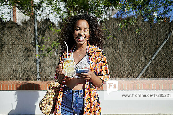 Lächelnde Frau mit Limonade und Smartphone an einem sonnigen Tag