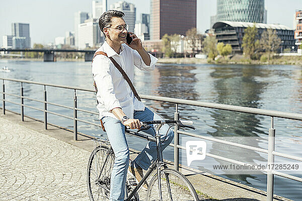 Lächelnder Geschäftsmann auf dem Fahrrad  während er am Fluss in der Stadt mit seinem Handy telefoniert