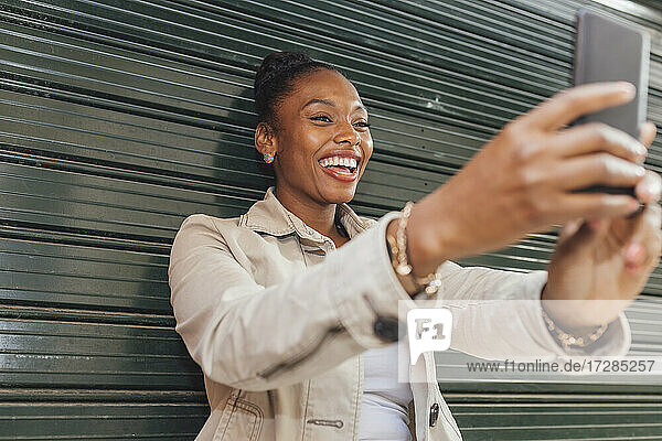 Fröhliche Frau  die ein Selfie mit ihrem Mobiltelefon macht  während sie vor dem Verschluss steht