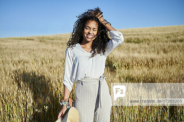 Lächelnde junge Frau steht mit der Hand im Haar in einem Weizenfeld an einem sonnigen Tag