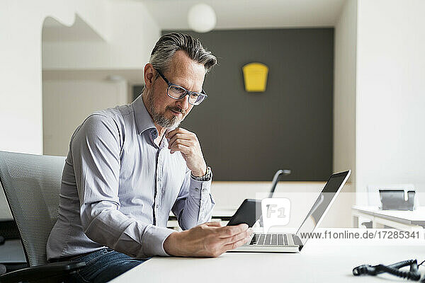 Männlicher Unternehmer  der am Schreibtisch sitzend ein Mobiltelefon benutzt