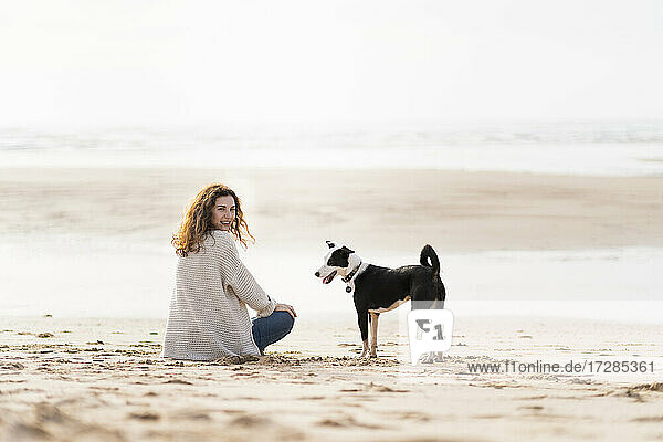 Woman looking over shoulder by dog while sitting on sand at beach during vacations
