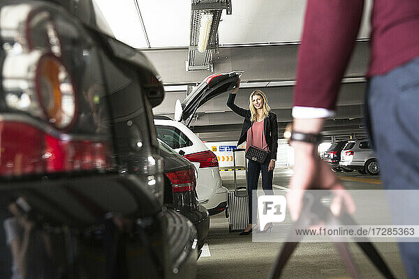 Lächelnde Frau  die den Kofferraum ihres Autos öffnet  während sie auf dem Flughafenparkplatz auf ihren Freund wartet