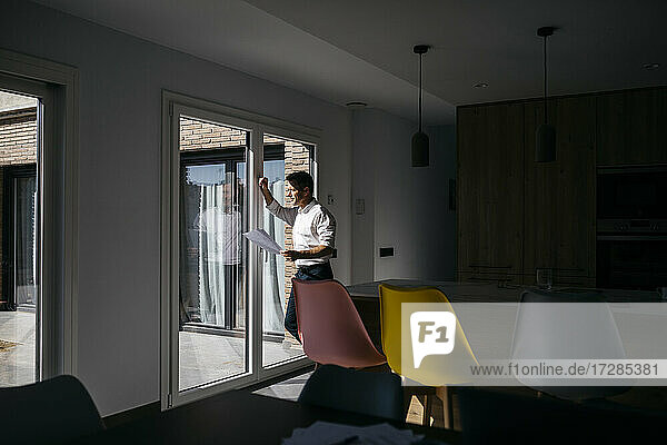 Male freelancer with documents looking through window at home