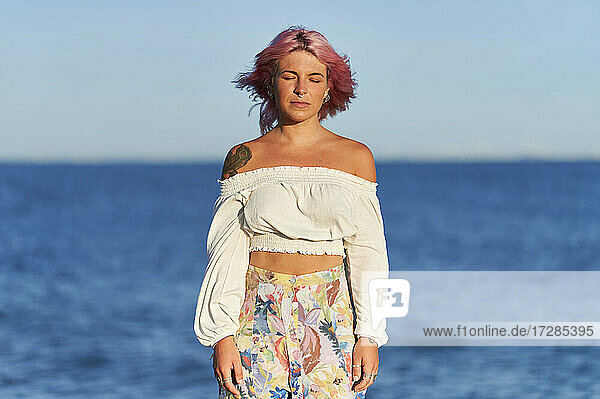 Junge Frau mit geschlossenen Augen steht vor dem Meer an einem sonnigen Tag