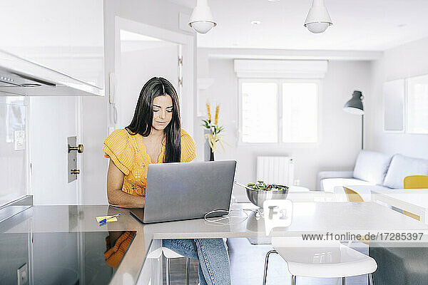 Frau benutzt Laptop  während sie neben einer Schüssel auf dem Küchentisch sitzt