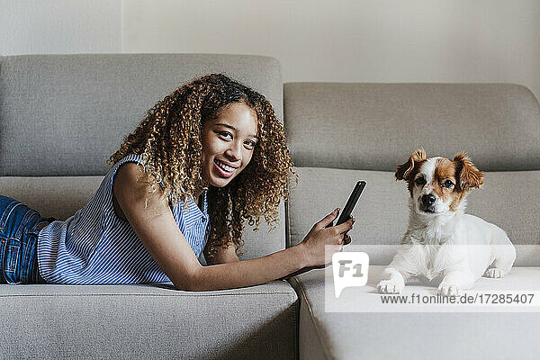 Curly haired woman with smart phone lying on sofa by dog in living room