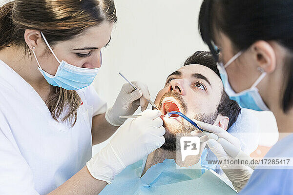 Zahnärztin mit Assistentin bei der Arbeit an den Zähnen eines männlichen Patienten in der Klinik während COVID-19
