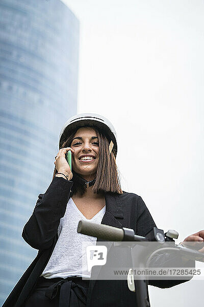 Junge Geschäftsfrau  die mit einem Mobiltelefon telefoniert  während sie mit einem Elektroroller steht