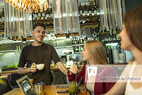 Lächelnder junger Mann mit Kaffeetasse im Gespräch mit Freundinnen am Tisch in einer Bar