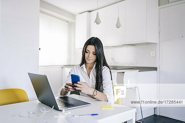 Junge schöne Frau  die ein Mobiltelefon benutzt  während sie zu Hause im Büro sitzt