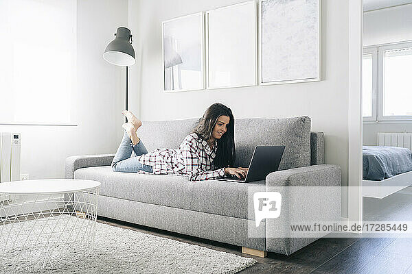 Smiling woman working on laptop while lying on sofa in living room