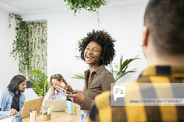 Fröhliche Geschäftsfrau mit digitalem Tablet  die einen männlichen Kollegen beim Diskutieren im Büro ansieht