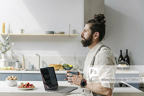 Thoughtful beard man holding coffee cup while sitting by laptop in kitchen