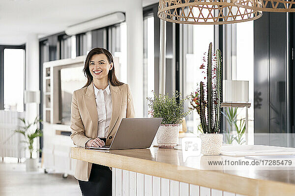 Smiling female entrepreneur looking away while standing in front of laptop at office cafeteria