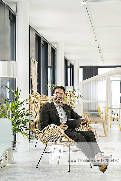 Geschäftsmann mit digitalem Tablet  der wegschaut  während er auf einem Stuhl in einer Büro-Cafeteria sitzt