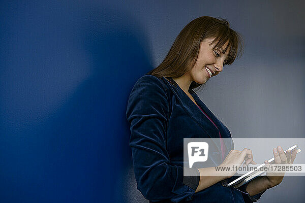Lächelnde junge Geschäftsfrau  die ein digitales Tablet an einer blauen Wand im Büro benutzt