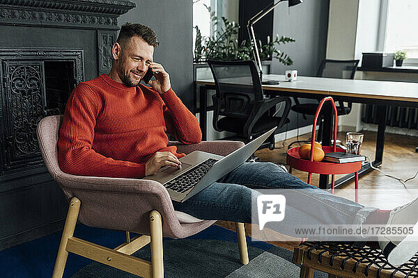 Männlicher Unternehmer  der einen Laptop benutzt  während er im Büro mit seinem Handy telefoniert