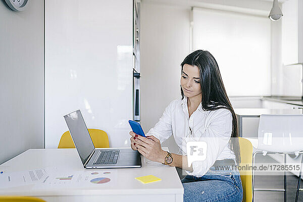 Junge Frau  die ein Mobiltelefon benutzt  während sie im Büro zu Hause am Laptop sitzt