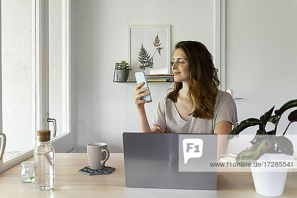 Lächelnde Frau  die ein Smartphone am Schreibtisch im Home Office benutzt
