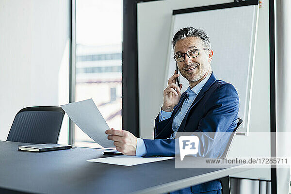 Mature male entrepreneur talking on smart phone sitting at desk looking away