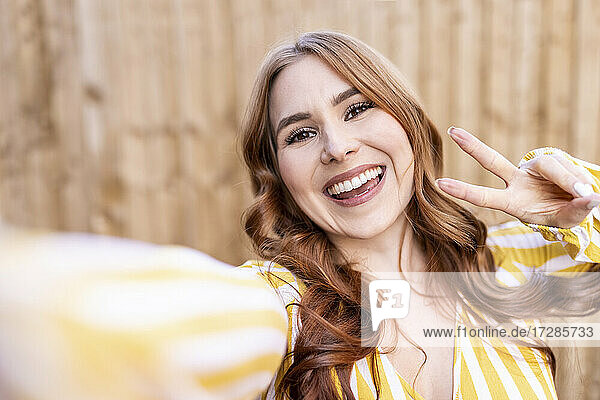 Cheerful redhead woman gesturing peace sign while standing against wooden wall