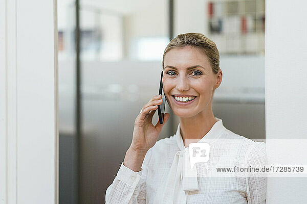Lächelnde blonde Geschäftsfrau  die mit einem Handy telefoniert und an der Tür eines Büros steht