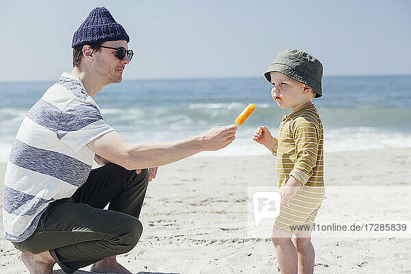 Mann mit Sonnenbrille füttert seinen Sohn am Strand mit Eiscreme