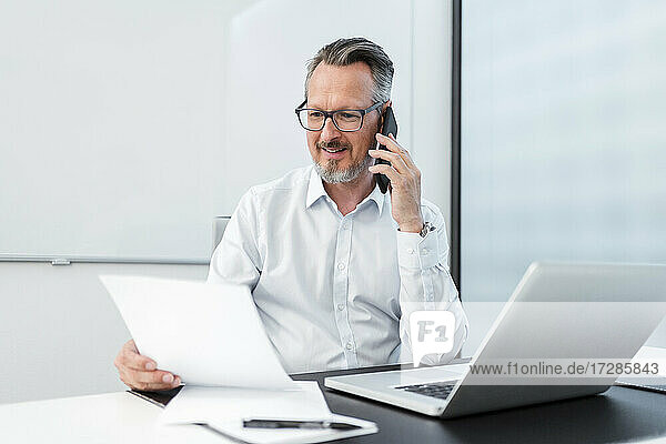 Businessman with document and laptop talking on mobile phone in office