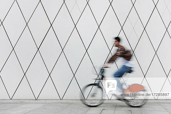 Young man cycling on footpath by wall