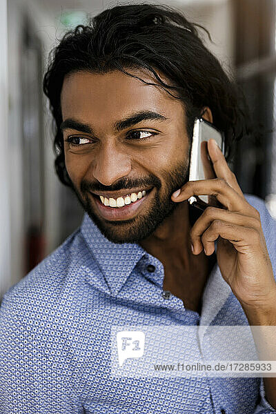 Lächelnder junger männlicher Unternehmer  der im Büro mit dem Handy telefoniert und wegschaut