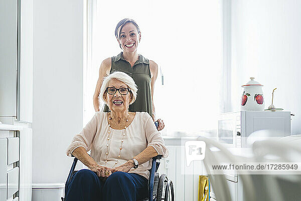Lächelnde Frau im mittleren Erwachsenenalter mit behinderter Großmutter  die auf einem Stuhl in der Küche zu Hause sitzt