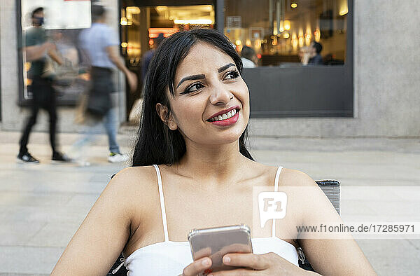 Lächelnde Frau träumt  während sie ihr Smartphone in einem Straßencafé hält