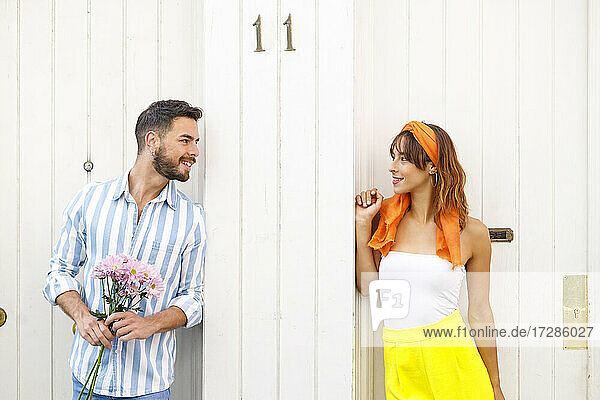 Junger Mann mit Blumenstrauß  der eine vor der Tür stehende Frau ansieht