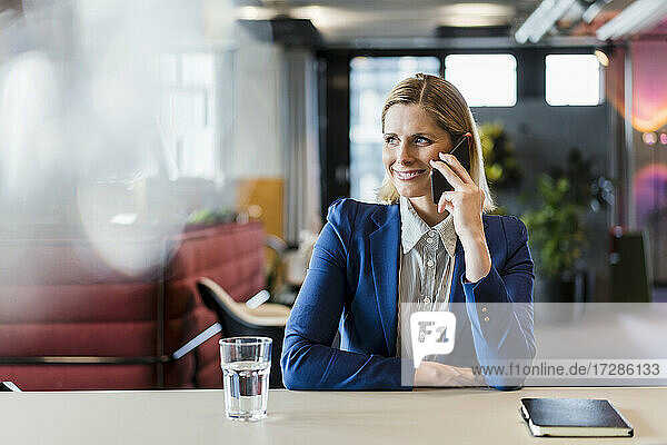 Lächelnde blonde Geschäftsfrau  die am Schreibtisch sitzend mit ihrem Handy telefoniert und wegschaut