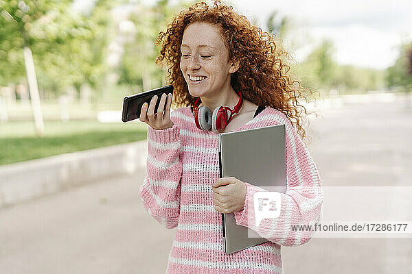 Smiling redhead woman carrying laptop while talking on smart phone through speaker at park