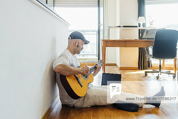 Männlicher Gitarrist spielt Gitarre am Laptop  während er zu Hause auf dem Boden sitzt