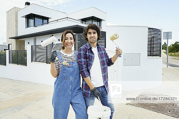 Lächelndes Paar mit Farbrolle vor einem neuen Haus