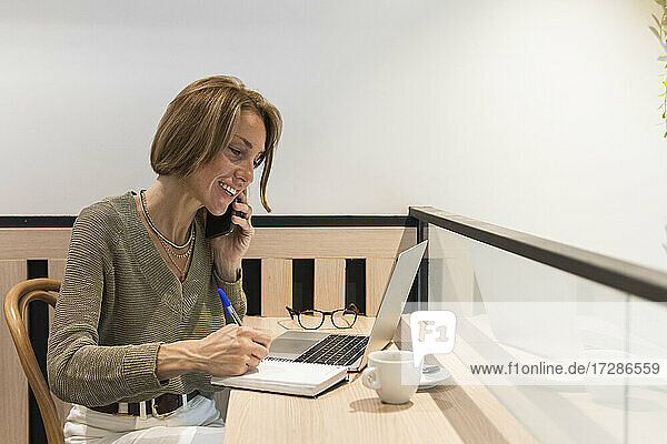 Frau mit Laptop  die in ein Buch schreibt  während sie in einem Café mit ihrem Handy telefoniert