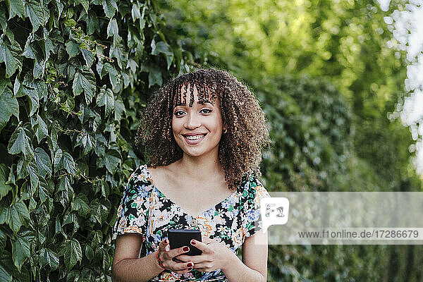 Smiling young woman holding smart phone while standing by ivy plant