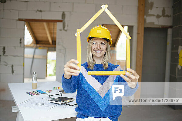 Lächelnde Bauunternehmerin  die ein hausförmiges gefaltetes Lineal in einer Wohnung untersucht