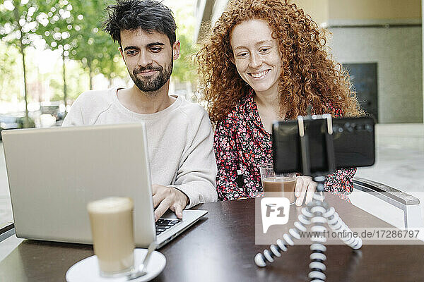 Lächelnde Freundin  die mit dem Handy filmt  während ihr Freund einen Laptop im Café benutzt