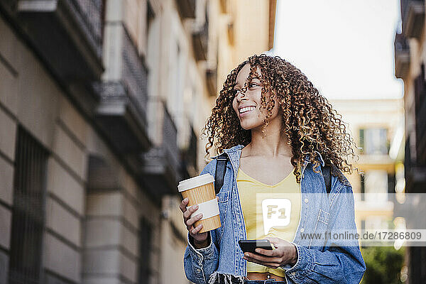 Beautiful woman looking away while holding reusable cup and smart phone in city